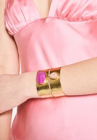 Un bracciale in metallo dorato presenta due pietre: una grande pietra rosa e una più piccola di un rosa chiaro, che contrastano con un vestito satinato rosa.