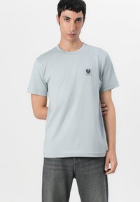 T-shirt de algodão azul claro com mangas curtas, decote redondo e um pequeno emblema preto na área do lado esquerdo do peito. Usado com jeans cinzentos.