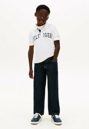Junge trägt ein weißes Polo-Shirt mit dem Schriftzug "HILFIGER", dunkle Jeans und marineblaue Sneakers, steht mit einer Hand in der Tasche vor weißem Hintergrund.