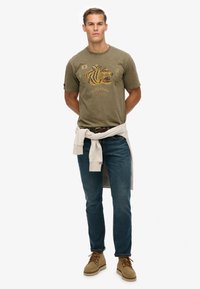Camiseta de algodón verde oliva con un gráfico de un tigre y texto, combinada con jeans azules y botas de gamuza beige; suéter de color claro atado a la cintura.