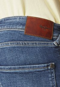 Indigo traperice s smešnim kožnim etiketom na struku na kojoj je utisnut natpis 'Pepe Jeans London'. Vidljivo šivanje i mali srebrni dugme.