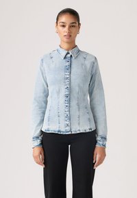 Γυναίκα με σκούρα μαλλιά σε κότσο, που φοράει ανοιχτό μπλε πουκάμισο από τζιν με κουμπιά και μαύρο παντελόνι, στέκεται μπροστά σε ουδέτερο φόντο.