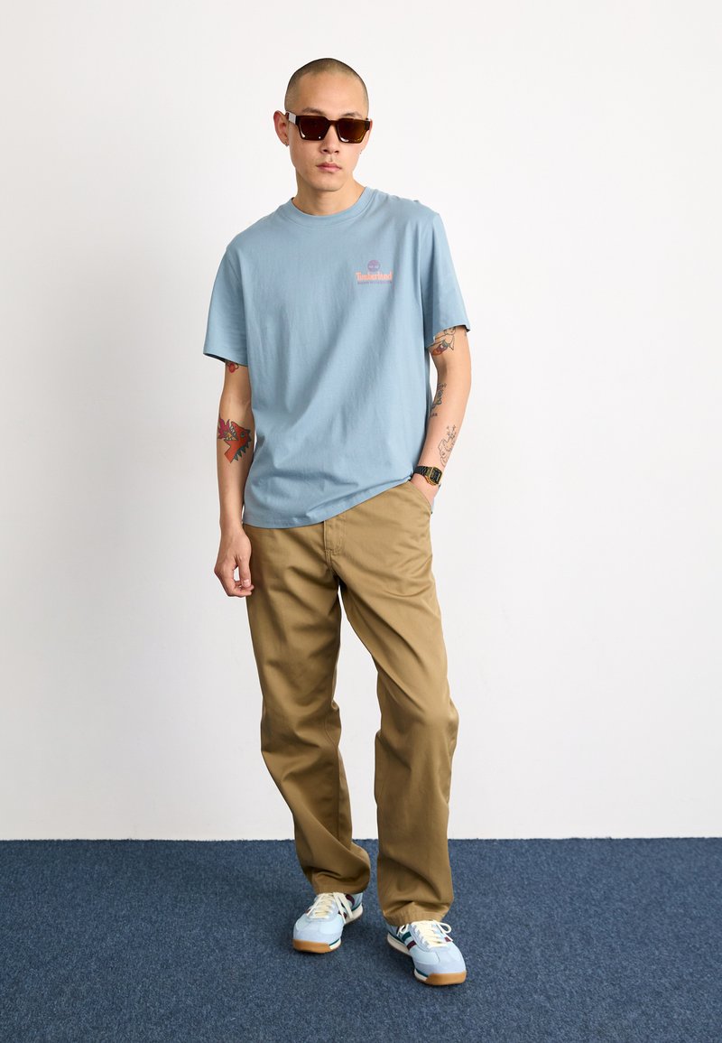 Camiseta de algodón azul claro con un logo impreso, combinada con pantalones khaki y zapatillas de colores. Las gafas de sol añaden un toque moderno.