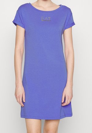 Mujer con vestido casual lila, de manga corta y largo hasta la rodilla, con logo dorado EA7 en el pecho, de pie frente a un fondo liso.