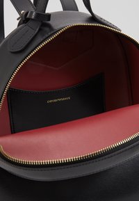 Mochila negra de cuero con una textura suave, con un interior rojo y detalles en cremallera dorados. Incluye un bolsillo interior con la marca.