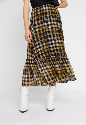 Kvinde iført en gul, sort og hvid ternet midi-nederdel med flæsedetalje og hvide ankelstøvler mod en ensfarvet baggrund.