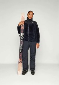 Fleece jacket in dark blue with a front zipper, paired with black ski pants and black ski boots. Individual holds pink and brown skis.