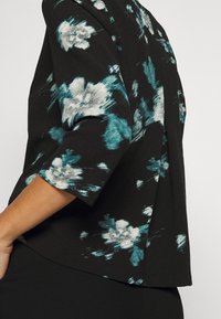 Sort top med teal og hvid blomstermønster, trekvartærmer, blødt stof, afslappet pasform, med en sideslids for ekstra bevægelse.