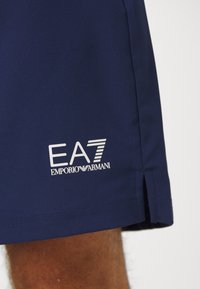 Tumši zili šorti, kas izgatavoti no gludas auduma, ar baltu EA7 Emporio Armani logo. Sānu izgriezums apakšējā malā.