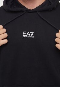 Svart hettegenser laget av bomullsmiks, med en liten hvit EA7-logo på brystet. Snor i hetten og teksturert stoff.