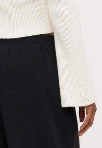Rippstrickpullover in Creme, kombiniert mit schwarzen, glatten Stoffhosen. Lange Ärmel mit weitem Bündchen und seitlichem Schlitzdetail.