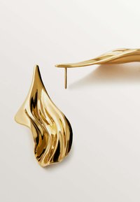 Pendientes de metal dorado con un diseño fluido y ondulante, que cuentan con un acabado brillante y un cierre en obispo para un uso seguro. Forma escultural con contornos elegantes.