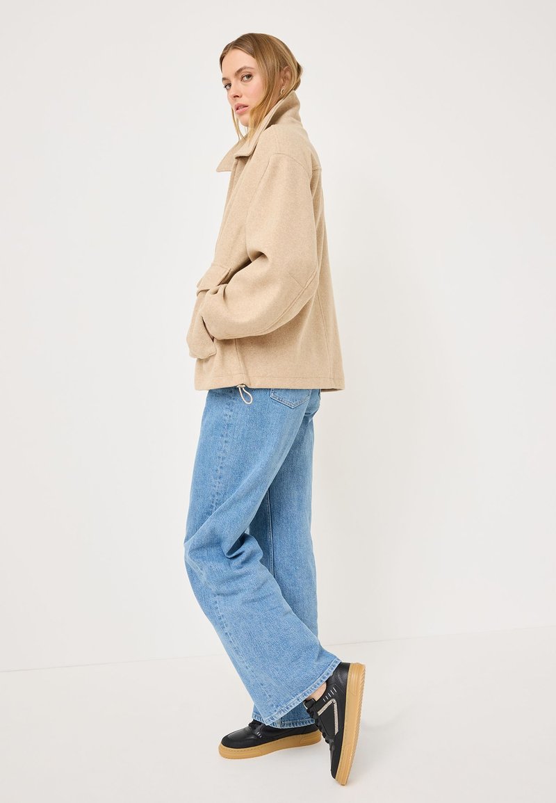 Giacca beige senza colletto in misto lana con spalle scese; abbinata a jeans azzurri chiari e larghi e sneakers nere con suole in gomma.