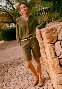 Femme portant une chemise à manches longues vert olive et un short, ceinture beige, sandales couleur camel, appuyée contre un mur en pierre en plein air avec des arbres et des feuilles de palmier.