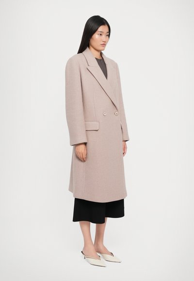 Woman standing in a beige double-breasted long coat, black wide-leg cropped pants, and white pointed kitten heels against a plain background.