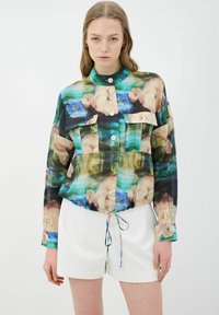 Blusa de estampado floral en tela transparente multicolor, con cuello alto, cierre de botones y dos bolsillos frontales. Bajo elástico con detalle de cordón.