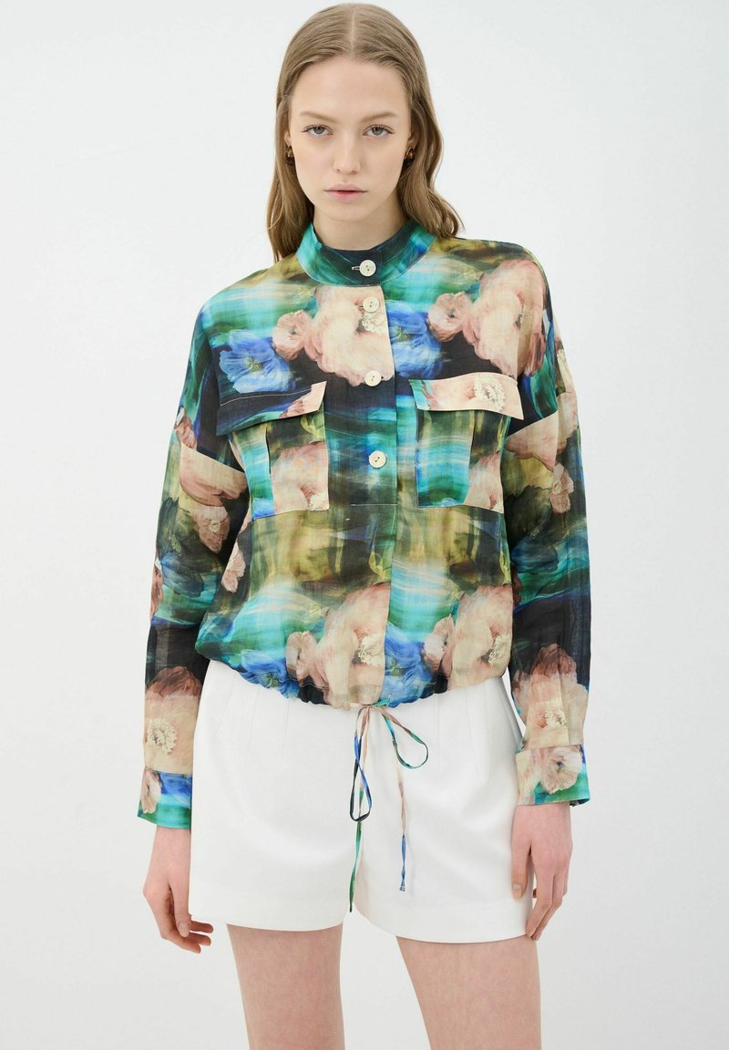 Blusa de estampado floral en tela transparente multicolor, con cuello alto, cierre de botones y dos bolsillos frontales. Bajo elástico con detalle de cordón.