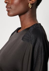 Μαύρη μπλούζα από ύφασμα με μαλακή υφή, με δαντελένιες και διχτυωτές λεπτομέρειες στον ώμο. Στρογγυλή λαιμόκοψη και άνετη εφαρμογή. Ορατό χρυσό σκουλαρίκι σε μορφή κρίκου.