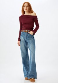 Bordeauxfarbenes, schulterfreies Oberteil mit langen Ärmeln, kombiniert mit hoch taillierten, hellblauen, ausgestellten Jeans und einem braunen Gürtel, stilvoll ergänzt durch braune Schuhe.