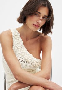 Robe en crochet blanc cassé avec une coupe à une épaule. Présente des appliqués floraux texturés le long de l'encolure. Tissu tricoté lisse.