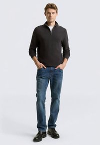 Dunkelgrauer Pullover mit Reißverschluss am Kragen, blaue Jeans und schwarze Schuhe. Schlanke Passform und strukturierter Stoff. Model steht mit Händen in den Taschen.