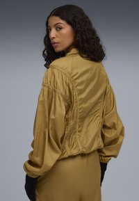 Giacca bomber in satin dorato con una vestibilità comoda, spalle arricciate e polsini a costine. Texture liscia e dettaglio del colletto alto.