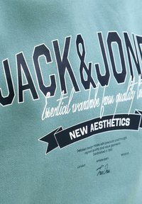 Felpa verde con scritta nera in grassetto "JACK & JONES." Include scritte bianche in corsivo e un banner nero con la scritta "NEW AESTHETICS." Tessuto dalla texture liscia.