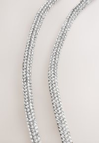 Zwei silberfarbene, mit Strass besetzte Stränge, die sich vor einem blassen beigen Hintergrund wölben.
