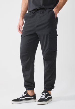Pantaloni cargo neri con vita elastica, tasche laterali e caviglie affusolate, realizzati in materiale resistente. Abbinati a sneaker nere e bianche.
