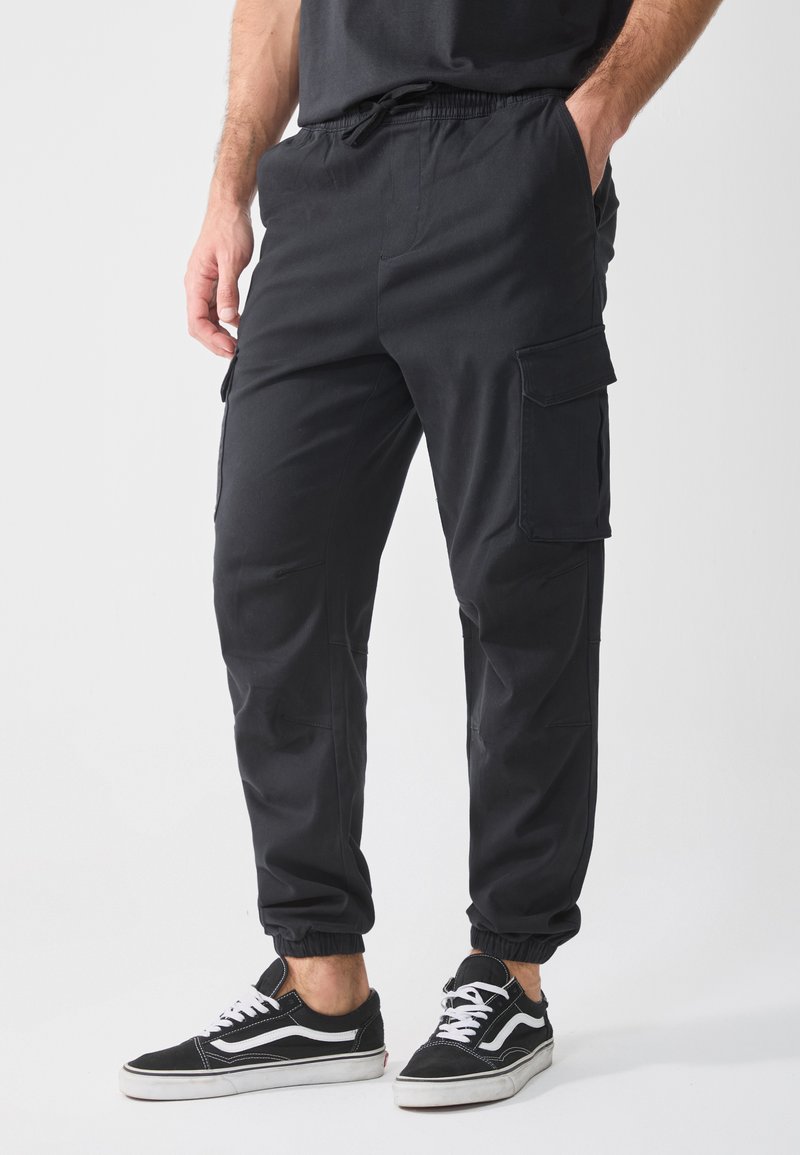 Schwarze Cargo-Hosen mit elastischem Bund, Seitentaschen und schmal zulaufendem Bein aus strapazierfähigem Material. Kombiniert mit schwarz-weißen Sneakers.