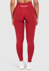 Leggings rossi a vita alta con una texture liscia, dotati di un dettaglio centrale a cucitura e un logo bianco del marchio in vita. Indossati con sneaker bianche.