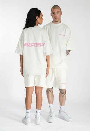 Übergroßes, hellcremefarbenes T-Shirt und passende Shorts mit pinkem "MULTIPLY"-Text und lässiger Passform; getragen mit weißen Sneakern und Socken.