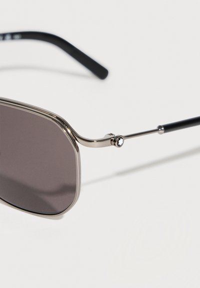 Κοντινή λήψη γυαλιών ηλίου με σκούρους ορθογώνιους φακούς, λεπτό ασημένιο μεταλλικό πλαίσιο και μαύρες άκρες στα κέρατα, σε λευκό φόντο.