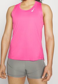 Žena v jasně růžovém sportovním topu bez rukávů a těsných šedých sportovních šortkách, stojící proti jednoduchému světlému pozadí.