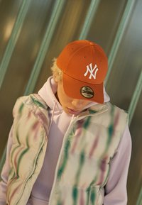 Oranger New York Yankees-Mütze mit weißem Logo, getragen mit einer pastelfarbenen, aufknöpfbaren Steppweste, die abstrakte Details in Grün, Pink und Weiß aufweist.