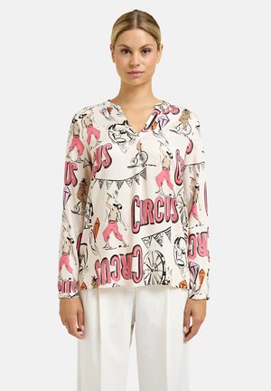 Frau trägt eine cremefarbene Bluse mit bunten, zirkusthematischen Illustrationen und Text, kombiniert mit weißen, weit geschnittenen Hosen.