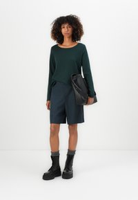 Woman with curly hair standing against white background wearing dark green long-sleeve top, navy shorts, black boots, gray socks, and holding a black bag.