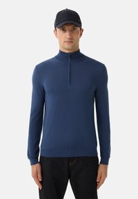 Mann in einem navyblauen Sweatshirt mit Viertelreißverschluss, schwarzer Baseballmütze und dunklen Jeans, der vor einem schlichten, hellgrauen Hintergrund steht.