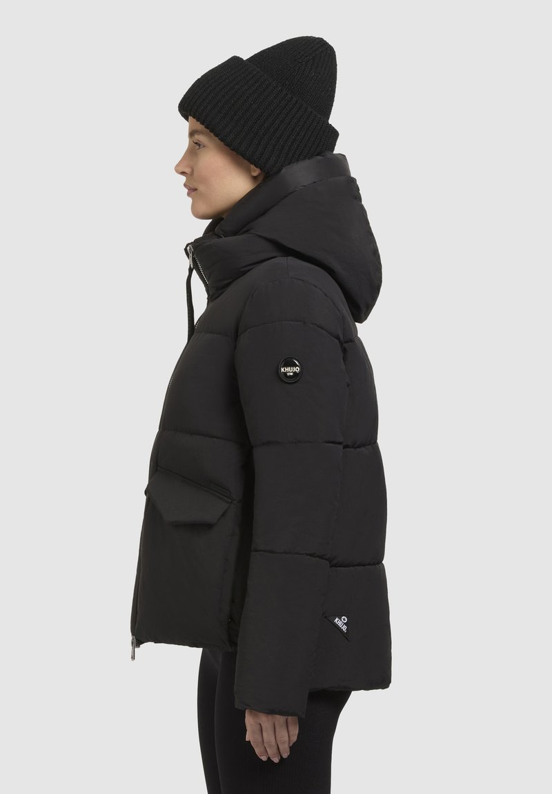 Schwarze Puffjacke mit Kapuze, mit genähten Abschnitten, einer auffälligen Fronttasche und einem Logopatch am Ärmel. Getragen mit einer schwarzen Mütze.