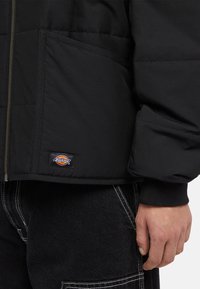 Černá péřová bunda se zapínáním na zip, s nášivkou loga blízko lemu a bočními kapsami. Tkanina vypadá hladce a je odolná.