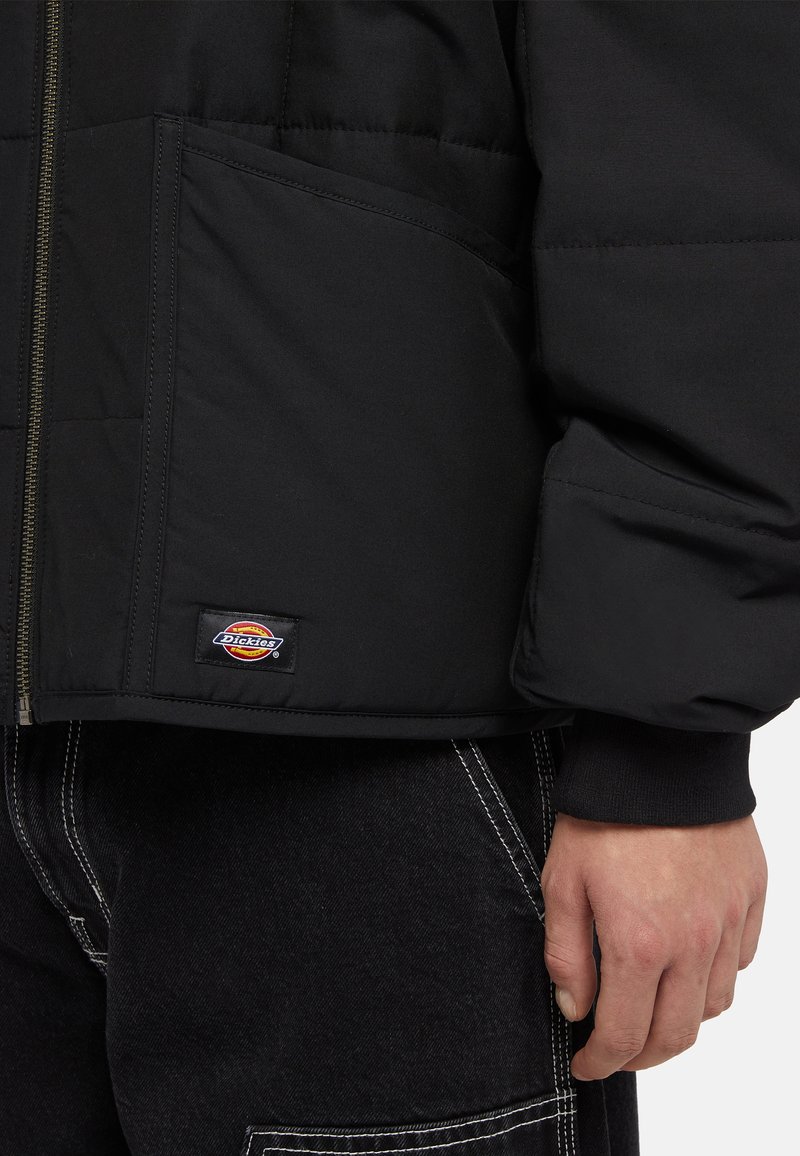 Doudoune noire avec fermeture éclair, présentant un logo en patch près de l'ourlet et des poches sur les côtés. Le tissu semble lisse et durable.