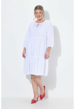 Donna sorridente che indossa un vestito bianco fino al ginocchio con maniche a tre quarti e scarpe rosse con tacchi alti, in piedi davanti a uno sfondo bianco.