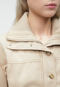 Kratki kadar osobe koja nosi bež jaknu s rebrastim, preklopnim kragnom, vidljivi su detalji gumba i patentnog zatvarača.