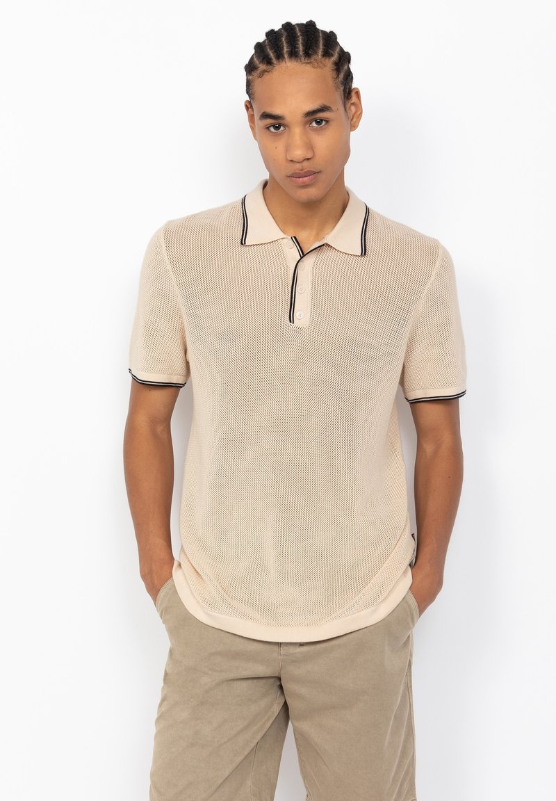 Polo beige avec un design tricoté texturé, présentant un col bordé de noir et une patte de boutonnage à deux boutons. Porté avec un pantalon de couleur claire.