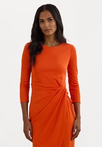 Vestido laranja de manga longa feito de tecido elástico, apresentando um detalhe de nó torcido na cintura e uma forma ajustada. Textura suave.