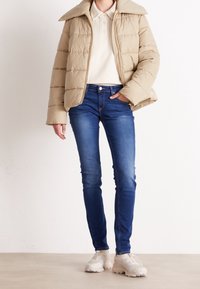 Beige puffer jacket with vertical seams, a large collar, and zipper closure. Paired with dark blue skinny jeans and light sneakers.