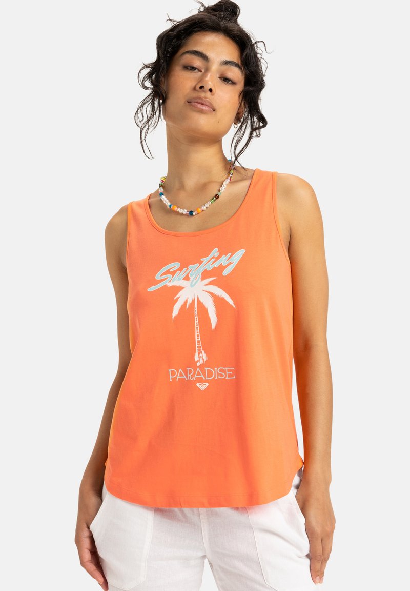 Kvinna klädd i en orange linne med texten "Surfing Paradise" och en palmträdsgrafik, som är kombinerad med vita byxor och ett färgglatt pärlhalsband.