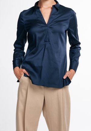 Femme portant un chemisier en satin bleu marine à manches longues avec un col en V et un pantalon beige à jambes larges, les mains dans les poches, debout devant un fond blanc.