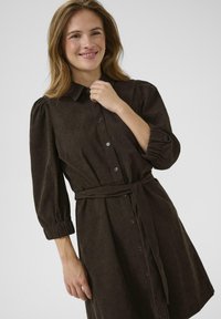 Robe-shirt en velours côtelé marron avec un col, des manches bouffantes, une fermeture à boutons sur le devant et une ceinture en tissu à la taille. Texture lisse.