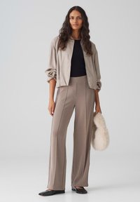 Beige bomberjack met gegatherde mouwen, zwarte aansluitende top, taupe wijde broek met zijnaad en zwarte flats, met een crèmekleurige nepbont clutch.